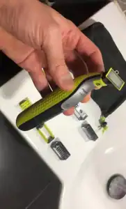 Afeitadora moderna en mano, accesorios de afeitado al fondo en superficie blanca.